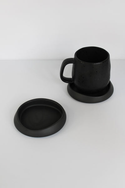 Modern stone-effect drink coasters in matte black with rounded design, shown with matching black ceramic mug